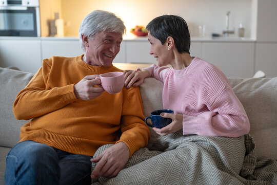 Senior husband and wife sit close together on couch holding tea cups, smiling elderly partners enjoy warm conversation, loving relationship, emotional connection and shared daily routine at home.