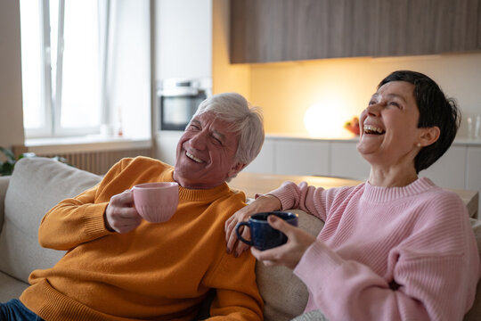 Happy elderly husband and wife lean back on couch and laugh together holding cups, affectionate senior spouses share humor, warm conversation and carefree leisure during retirement at home.