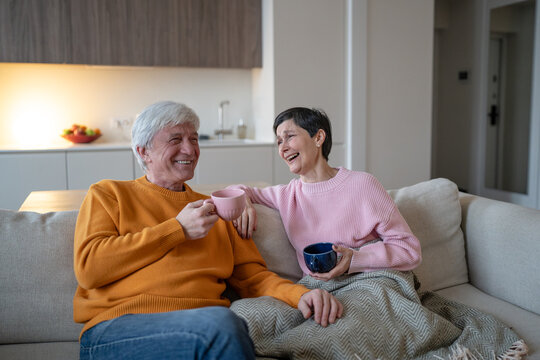 Happy senior married couple sits on couch holding colorful cups and laughs during conversation, joyful husband and wife share tea, humor, companionship and relaxed retirement time together.