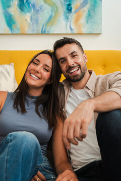 Warm candid portrait showing a joyful couple resting close on a bright couch, grinning at the camera with relaxed posture and natural smiles. The scene conveys affection, companionship, and everyday