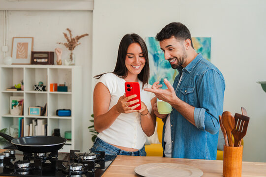 A cheerful couple shares a spontaneous kitchen moment as the woman displays a bright red smartphone to her partner. They exchange amused glances while exploring apps and footage together. Warm moment.