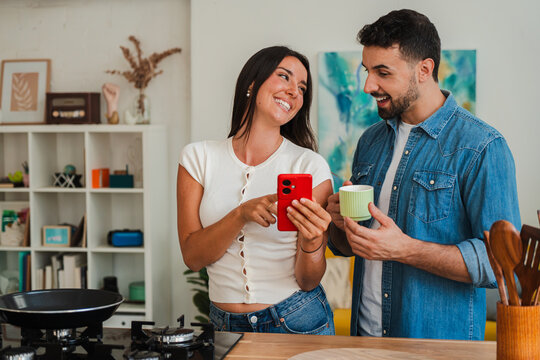 A couple engages in a light moment with a red smartphone while standing at a kitchen counter, sharing a quick glance and a cup of beverages. The scene conveys everyday connection and collaboration in