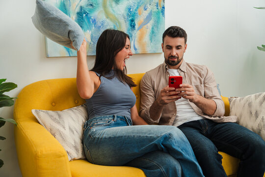 Two people share a cozy moment on a bright sofa as one taps a phone and points to the screen, while the other leans closer with a smile, creating a warm, relaxed, partner activity mood. Warm vibe now.