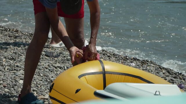 Inflatable boat pebble beach, man deflating yellow rubber dinghy on sunny shore by the sea