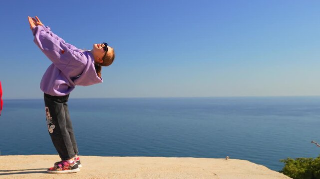 Talented gymnast performs an acrobatic backbend and handstand sequence on a coastal cliff edge. Static wide shot.