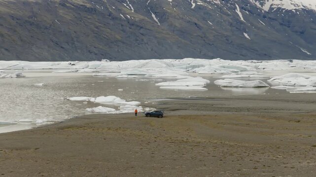 Fjallsarlon glacier lagoon with an SUV, person in red jacket, floating ice and Fjallsjokull glacier beneath snow covered mountains in South Iceland, drone establishing shot