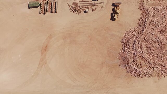 Top-down drone footage showing heavy machinery working at a construction site, with a yellow wheel loader moving gravel and a dump truck leaving the area, revealing tire tracks in the sand