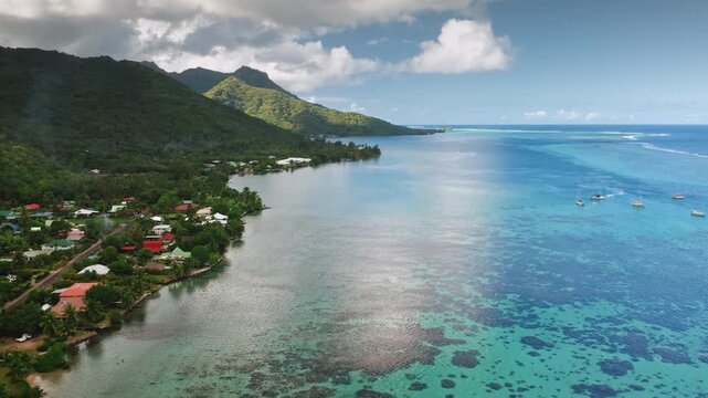 Moorea tropical coastline, lush green mountains, coastal village along turquoise lagoon revealing coral reefs, boats cruising calm Pacific water under clear blue sky. Aerial French Polynesia landscape