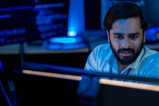 Worried programmer without glasses gaping at his monitor with raised brows, a close-up of shock at an unexpected result in a dark office lit by blue screens and lamps