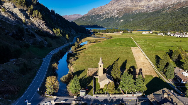 Aerial view of the historic Church of San Lorenzo surrounded by lush green meadows, a winding river, and a mountain road in Sils im EngadinSegl, Grisons, Switzerland.