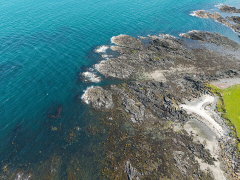 Aerial view of a rocky coastline with dark jagged stones meeting the deep blue sea and a patch of green grass in Donaghadee, Northern Ireland, United Kingdom.