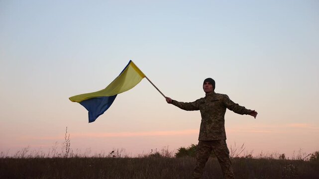 Male military waving blue-yellow banner on liberated territory occupied by russia. Young soldier of ukrainian army lifted flag of Ukraine in honor of victory against russian aggression at dusk