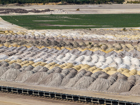 Aerial view of Tagebau Welzow-Sud open-pit mine with rows of textured sand and soil mounds in white, yellow, and grey, conveyor belt, and green fields in Spremberg, Brandenburg, Germany.