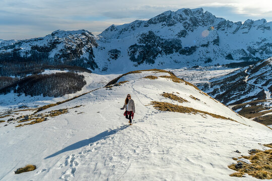 Female traveler hiking across a snow-covered mountain ridge surrounded by dramatic peaks and winter scenery in Montenegro. Outdoor adventure, trekking, active vacation and mountain tourism concept.