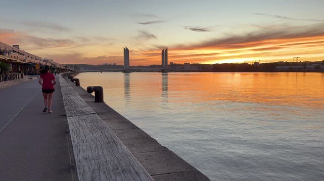 Static shot of sunrise reflections in the Garonne River in Bordeaux, with the Chaban-Delmas Bridge in the background and the Bacalan Quay in the foreground with a runner, a cyclist and a scooter