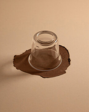 Clear glass cup placed upside down on a dark brown circular puddle of liquid on a light beige surface creating a simple minimalist composition