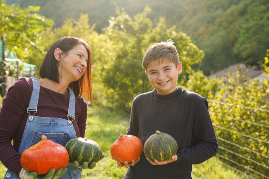 Happy mother and teenager son having fun harvesting organic pumpking during fall season at house garden - Farmer lifestyle, family and autumn season concept - UGC content