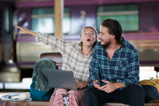 Young couple traveling, surprised and pointing at something exciting at a train station, planning a new journey with a laptop and smartphone