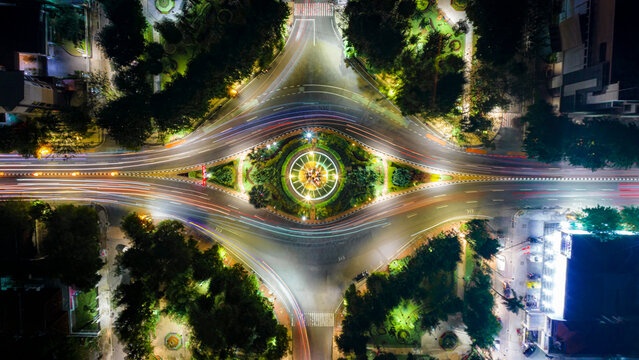 Aerial view of Monumen Bambu Runcing roundabout at night with vibrant light trails from traffic and illuminated greenery in Kecamatan Genteng, Jawa Timur, Indonesia.