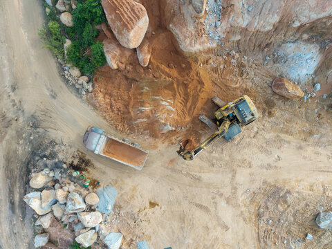 Aerial view of an excavator loading a dump truck at a mining site with piles of earth and rocky terrain in Bukit Tambun, Penang, Malaysia.