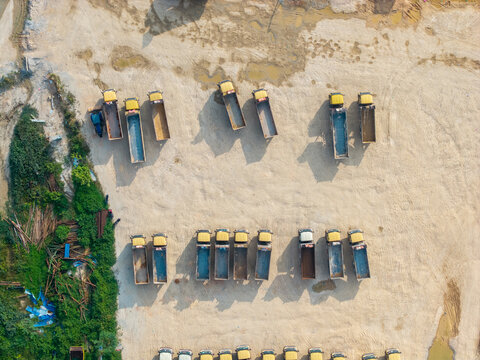 Aerial view of a construction area with rows of yellow dump trucks parked on a dirt lot near green vegetation in Bukit Tambun, Penang, Malaysia.