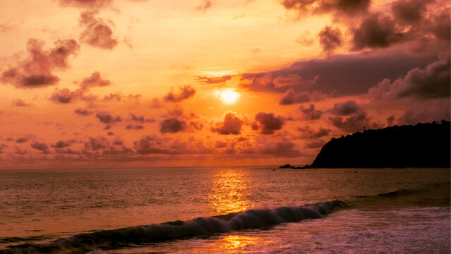 Aerial view of Modangan Beach at sunset with golden light reflecting on the ocean waves, dramatic clouds, and a dark headland silhouette in Donomulyo, East Java, Indonesia.