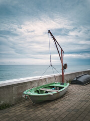 Small green fishing boat with a mechanical hoist winch on a coastal promenade by the ocean under cloudy sky. Maritime transport equipment on a concrete promenade on a overcast day. © Wizarto