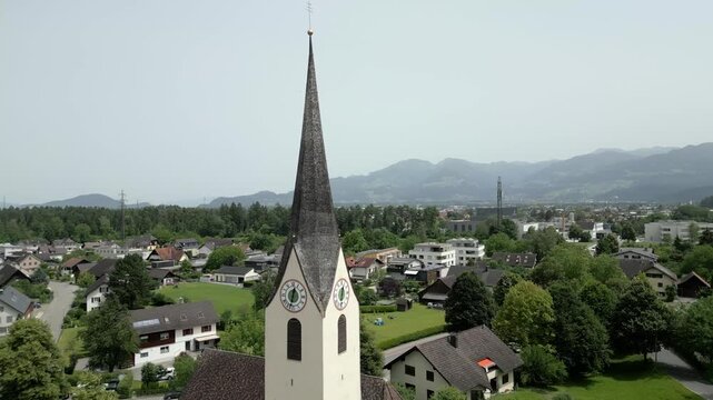 Aerial view of a church steeple rising above a small town with lush green trees and mountains in the background in Liechtenstein.