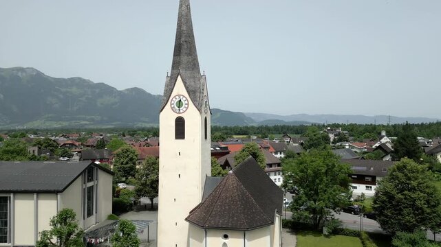 Aerial view of a historic church with a tall steeple and tiled roof, surrounded by a cemetery and green lawns in Liechtenstein.