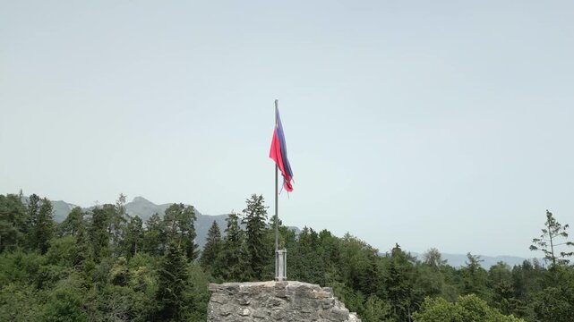 Aerial view of the national flag of Liechtenstein waving atop a historic stone fortification surrounded by dense forest and mountain landscape in Liechtenstein.