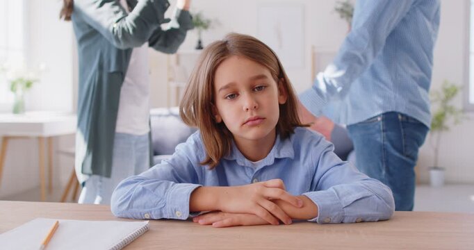 Frustrated upset child boy sitting at desk while his mother and father quarrelling and arguing at home. Divorce, psychological trauma and family conflicts concept. 4k video. Slow motion.