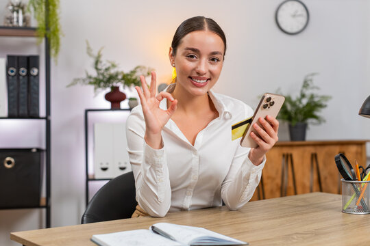 Young businesswoman at home office checks price on smartphone enters card confirms payment and feels relief. Freelancer girl at table shows ok sign writes expense for budget and saves order id today.