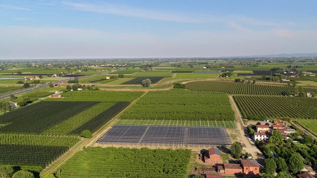 Aerial view of solar panels installed in agricultural fields with farm buildings and lush green crops in the countryside of Faenza, Emilia-Romagna, Italy.