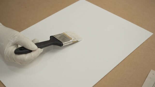 Gloved hand using a paintbrush to apply white paint or primer to a sheet of paper or board