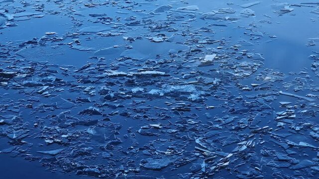 Thin ice melting on water in early spring. Texture of broken ice on river surface, close-up. Ice fragments in blue water during thaw. Blue water with small ice close-up. Thin winter ice on water