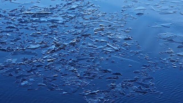 Thin ice melting on water in early spring. Texture of broken ice on river surface, close-up. Ice fragments in blue water during thaw. Blue water with small ice close-up. Thin winter ice on water