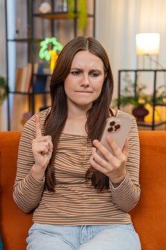 Young woman at home sees app error on smartphone frowns shakes finger no no then taps retry. Caucasian girl on sofa feels tense checks card and tries again hopes fix will work and restore access back