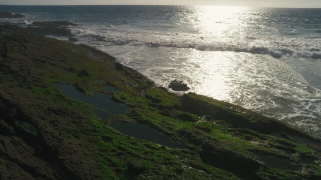 A slow upward tilt reveals waves washing beside algae-covered rocks and shallow pools. Bright reflections give the rugged coast an atmospheric feel for nature and ecology content.