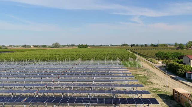 Aerial view of a large metal framework for a solar power plant in a rural field with green trees under a clear blue sky in Faenza, Emilia-Romagna, Italy.