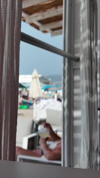 Vertical view from the shaded interior of the gazebo onto a sunny, sandy beach with thatched umbrellas, deck chairs and people relaxing in the blurry background. Empty space for text.
