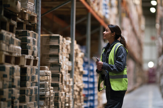 A woman in a safety vest inspects stock on high shelves in a warehouse, demonstrating her role in ensuring product availability and effective inventory management.