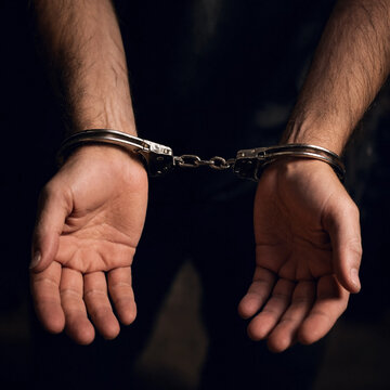 Adult man wearing metal handcuffs holds his hands forward against a dark background, suggesting arrest, detention, or criminal justice.