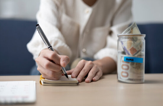 A young woman is sitting and shopping for a house using money she saved in a piggy bank, while noting down expenses and keeping track of her finances.