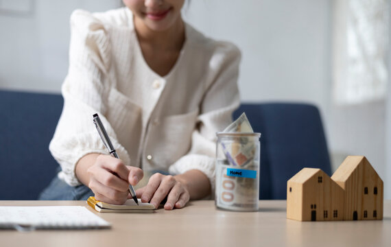 A young woman is sitting and shopping for a house using money she saved in a piggy bank, while noting down expenses and keeping track of her finances.