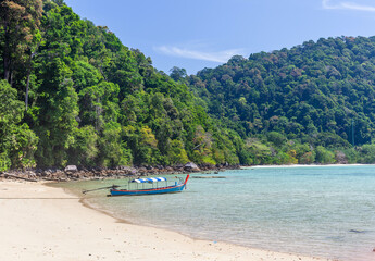 Fototapeta premium A longtail boat rests on a white sand beach beside clear turquoise water at Koh Surin, Thailand, with lush green hills under a blue sky.