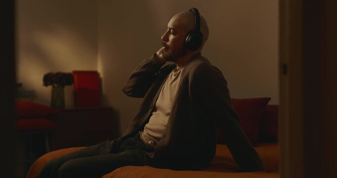 Bald man wearing headphones touching ear cup sitting on bed in cozy warm bedroom, listening to music or podcast with eyes closed, serene evening indoor relaxation atmosphere, lifestyle side view.   