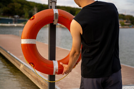 Man taking orange lifebuoy from stand on wooden pier at lake shore, representing water safety, lifeguard assistance, emergency response, and rescue equipment outdoors.