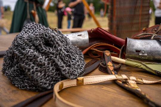 Medieval knight armor elements, steel chainmail coif, metal bracers, and leather belts with brass fittings on a wooden table at historical reenactment festival.