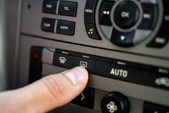 Driver finger pressing rear window defroster button on car climate control center console panel, turning on heated glass system to clear view.