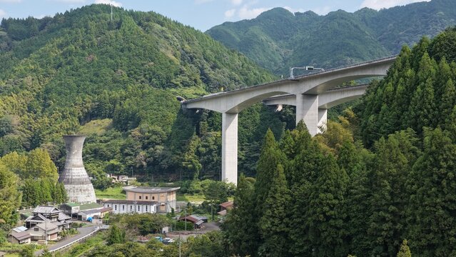 Aerial view of a large concrete viaduct bridge spanning a lush green valley with a small village and an industrial tower nestled among the forested mountains in Shizuoka, Shizuoka, Japan.